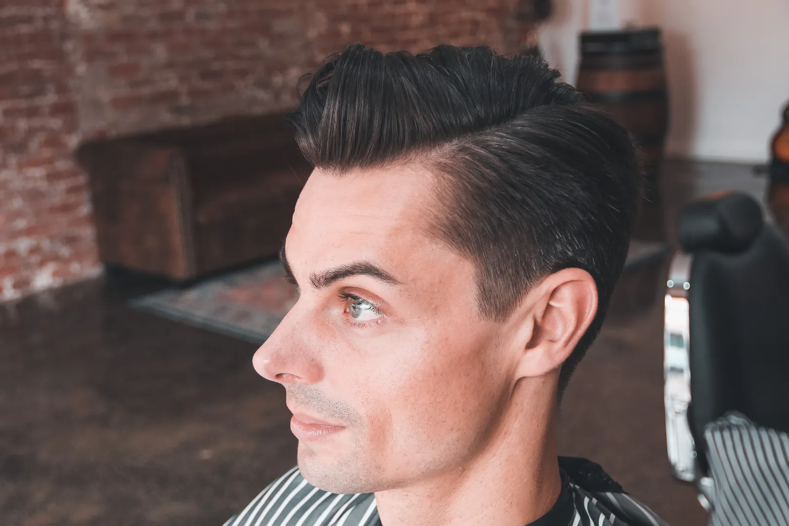 A finished high pompadour with a tight skin fade, shot in profile against the blurred exposed-brick wall at Amunra.