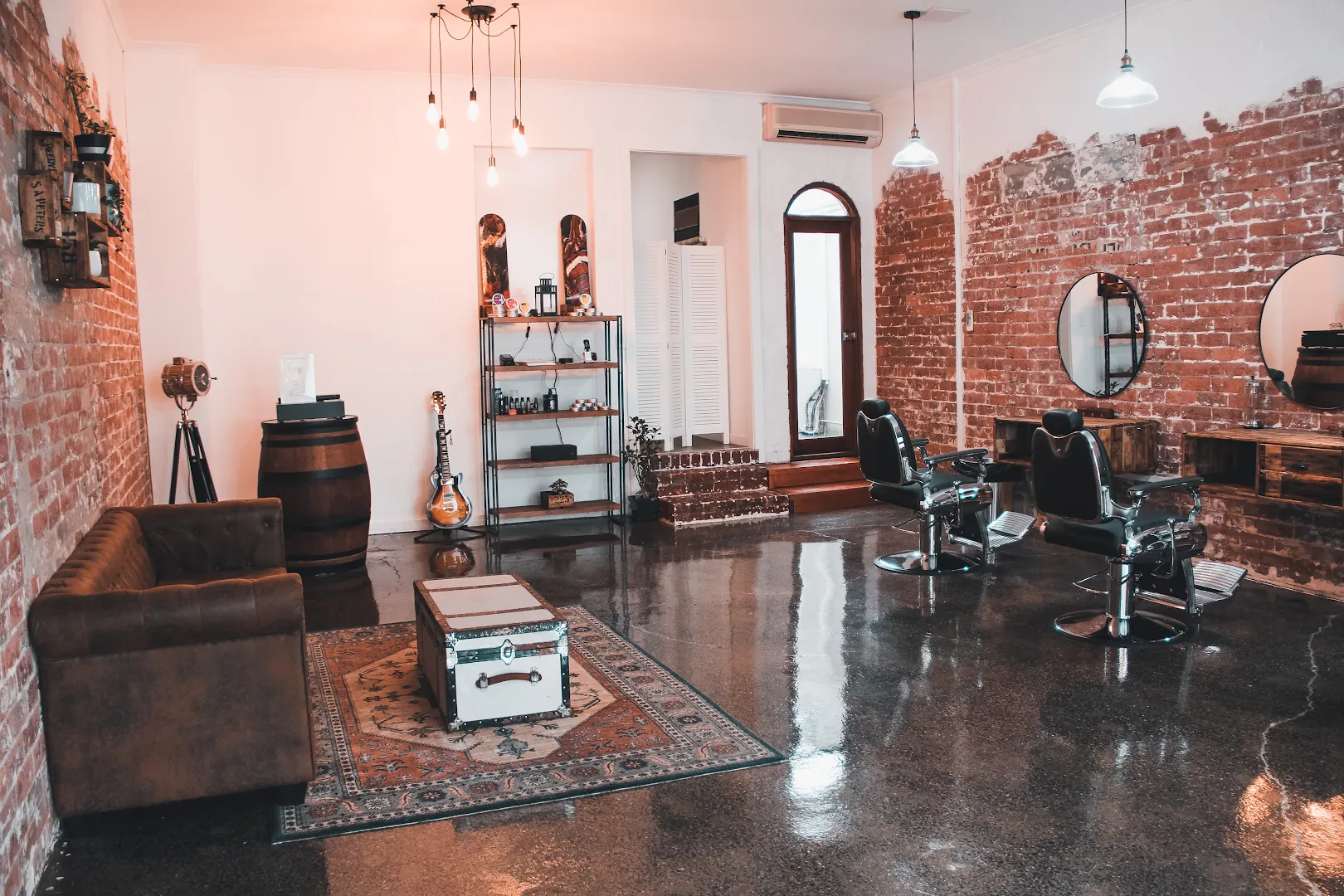 Wide view of Amunra's interior — two chrome barber chairs against exposed red brick, a leather Chesterfield and steamer trunk in the foreground under an Edison-bulb cluster.